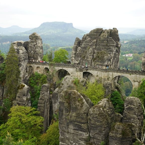 Historische Steinbrücke verbindet hohe Sandsteinfelsen, umgeben von grünem Wald mit fernen Bergketten im Hintergrund.