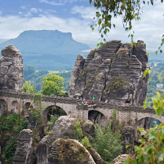 Historische Steinbrücke zwischen dramatischen Sandsteinfelsen mit Besuchern, umgeben von grünem Wald und Bergblick im Hintergrund.