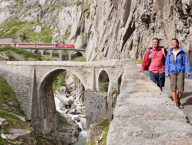 Zwei Wanderer wandern an einer Steinmauer entlang, im Hintergrund fährt ein Zug