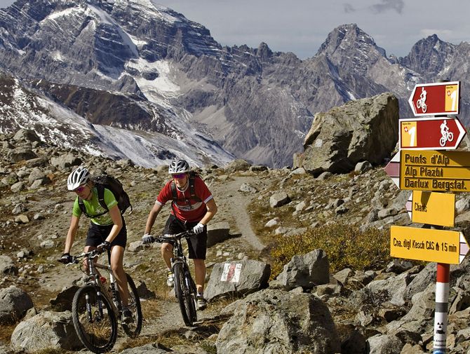 Zwei Mountainbiker in den Alpen. Ein Wegweiser zeigt ihnen die weitere Route zum Kesch in den Bündner Bergen.