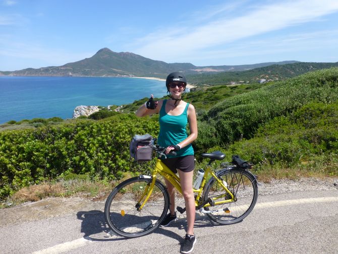 Dana steht mit ihrem Velo auf einer Strasse mit Sicht auf eine Bucht auf Sardinien.