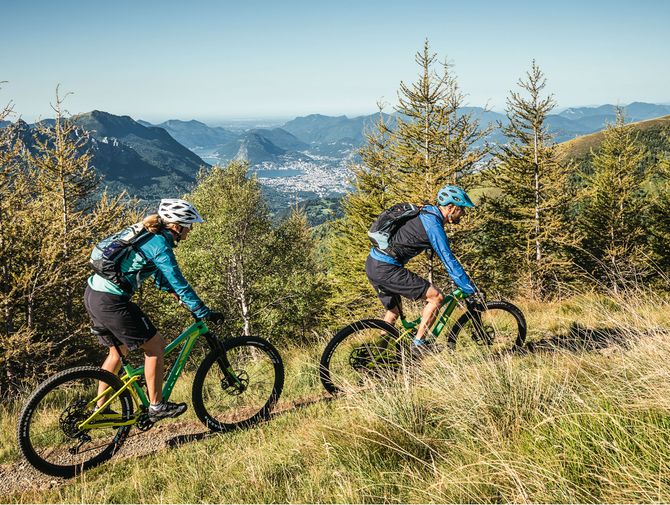 Ein Mountainbike Pärchen fährt über eine Wiese. In der Mitte liegt im Tal, umgeben von Bergen, liegt Lugano.