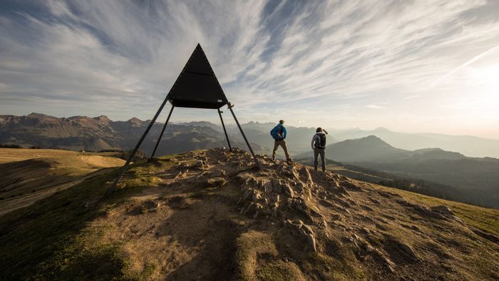 Zwei Wanderer stehen auf einer Bergspitze und blicken in die Berglandschaft vor Ihnen. Neben den Wanderern steht ein Metalliges Dreieck dessen Spitz sich in den Himmel richtet.