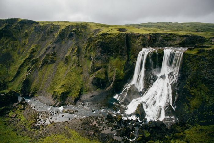 Das ganze Bild ist von eine grünen Klippe gefüllt von der auf der rechten Seite ein grosser Wasserfall herunter fliesst. Der Himmel ist grau und bewölkt.