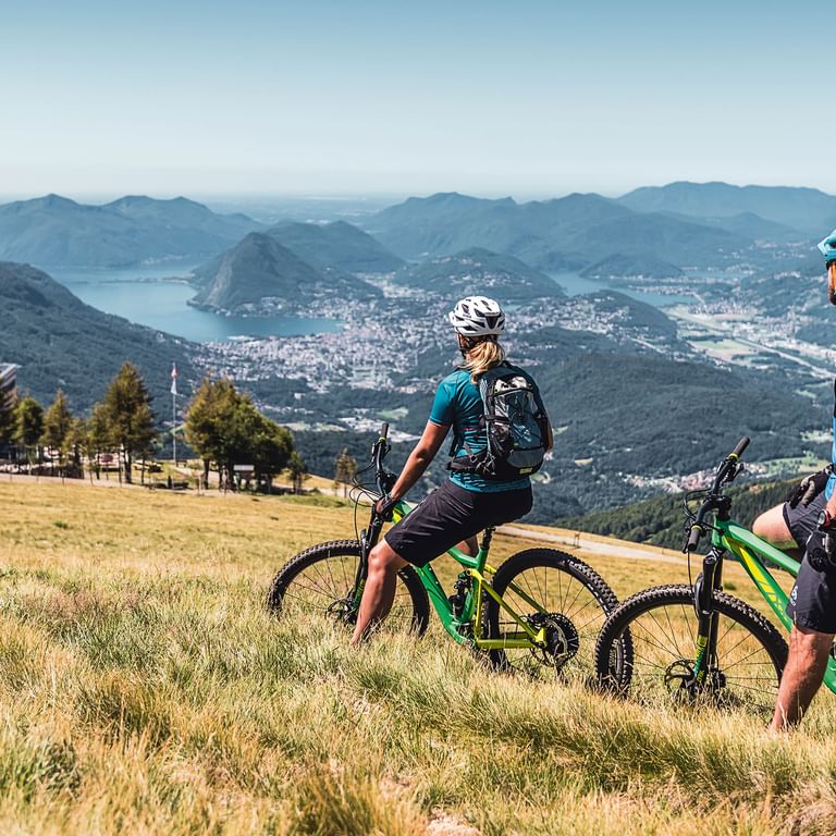 Zwei Mountainbiker mit Helmen und Rucksäcken stehen in einer goldenen Wiese und blicken auf Lugano mit See und Bergen.