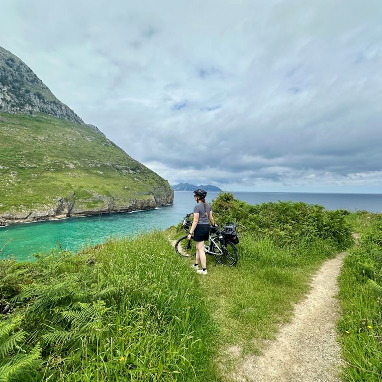 Cyclist with packed bike on gravel path overlooking turquoise bay at Parque de Sonabia. Green meadows, rocky cliffs, and ocean under cloudy sky.