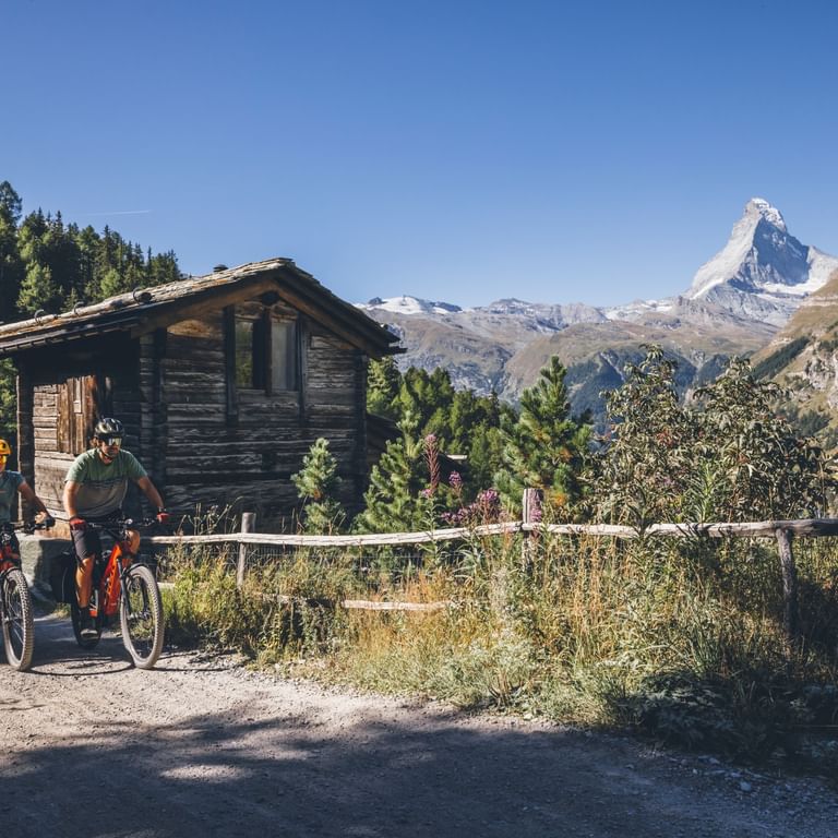 Zwei Radfahrer mit Rädern auf einer Schotterstraße neben einem Holzchalet, mit dem ikonischen Matterhorn im Hintergrund unter klarem blauen Himmel.