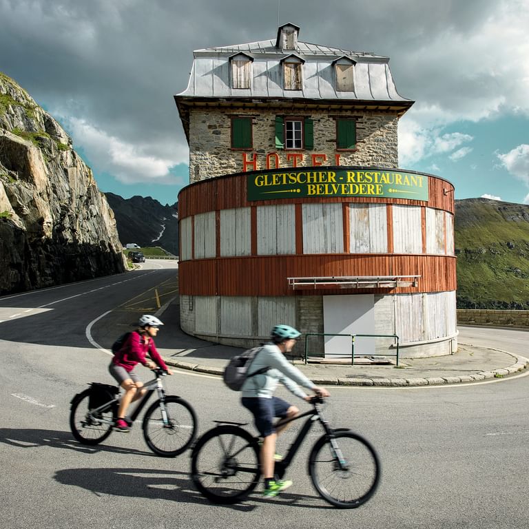 Zwei Radfahrer fahren am markanten runden Hotel Belvedere-Gebäude auf dem Furkapass vorbei, mit dramatischen Felsklippen und Berghängen.