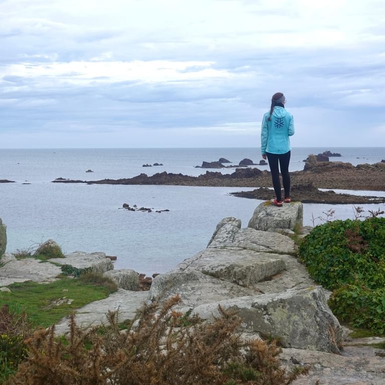 Frau in türkisfarbener Jacke steht auf Küstenfelsen bei Plougrescant, Bretagne, mit Blick auf das Meer und verstreute Felsformationen.