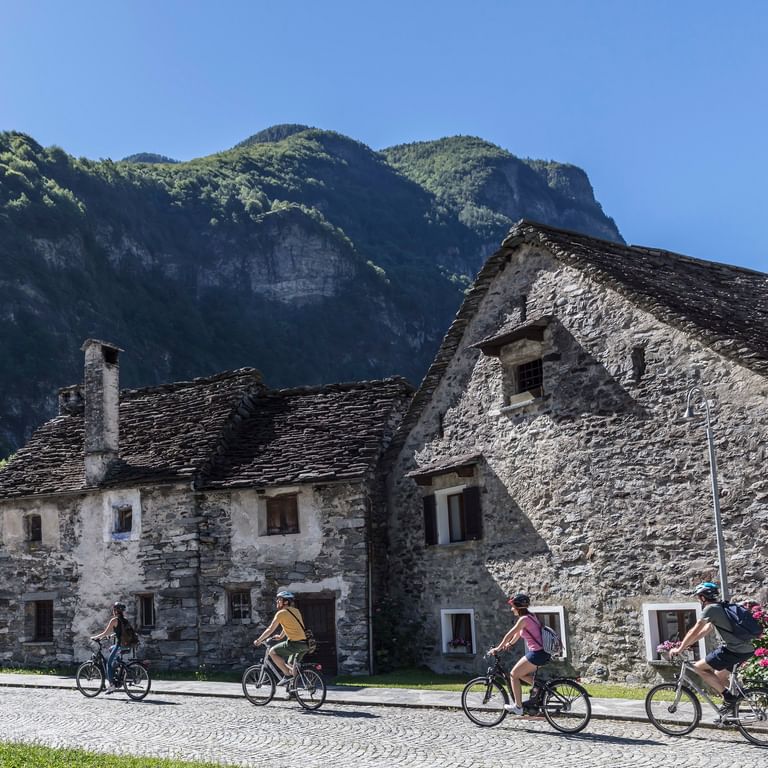 Gruppe von Radfahrern fährt durch traditionelles Steindorf in Vallemaggia, Tessin, mit historischen Gebäuden und grünen Bergen im Hintergrund.