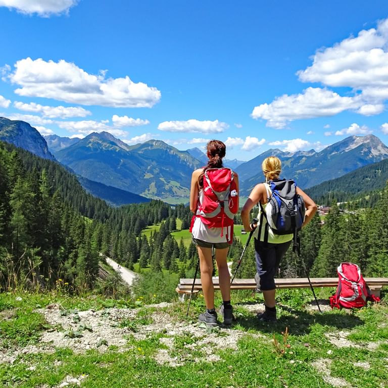 Zwei Wanderer mit Rucksäcken stehen an Holzgeländer und blicken über Alpental mit bewaldeten Bergen und blauem Himmel mit weißen Wolken.