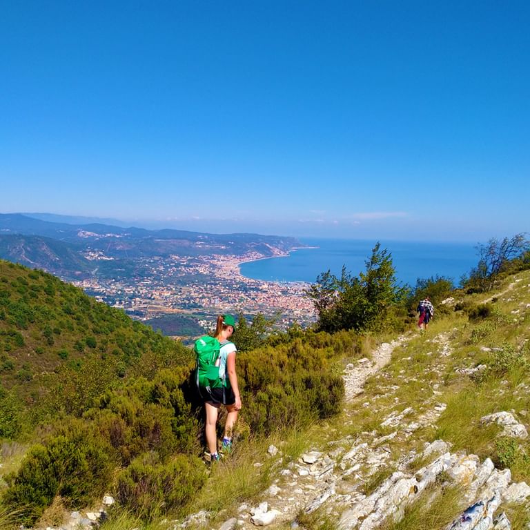 Two hikers with backpacks walking on a rocky mountain trail overlooking the Ligurian coast with blue sea and coastal towns below.