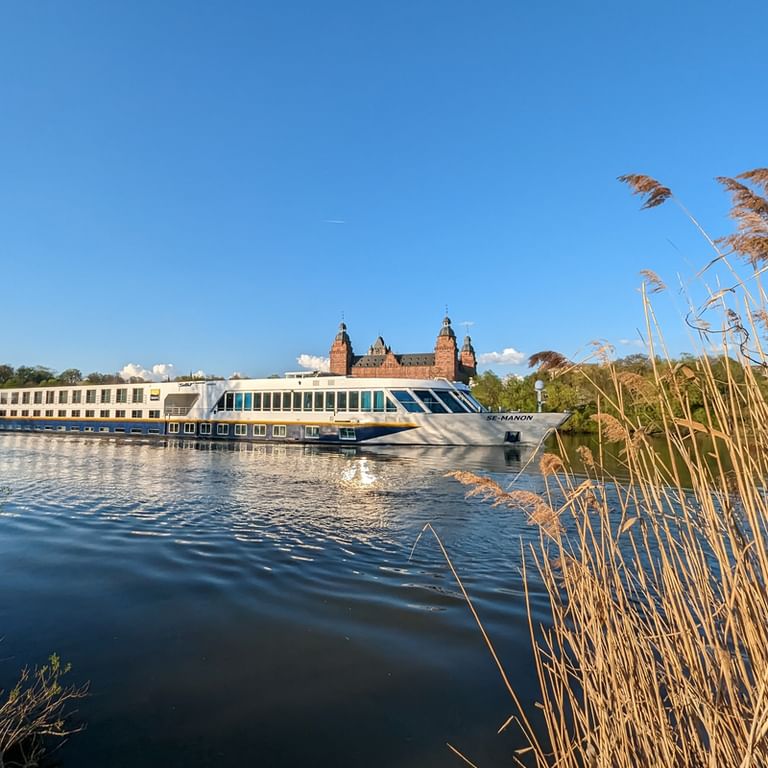 MS SE-MANON river cruise ship sailing on calm water with golden reeds in foreground and historic castle with towers in background under blue sky.