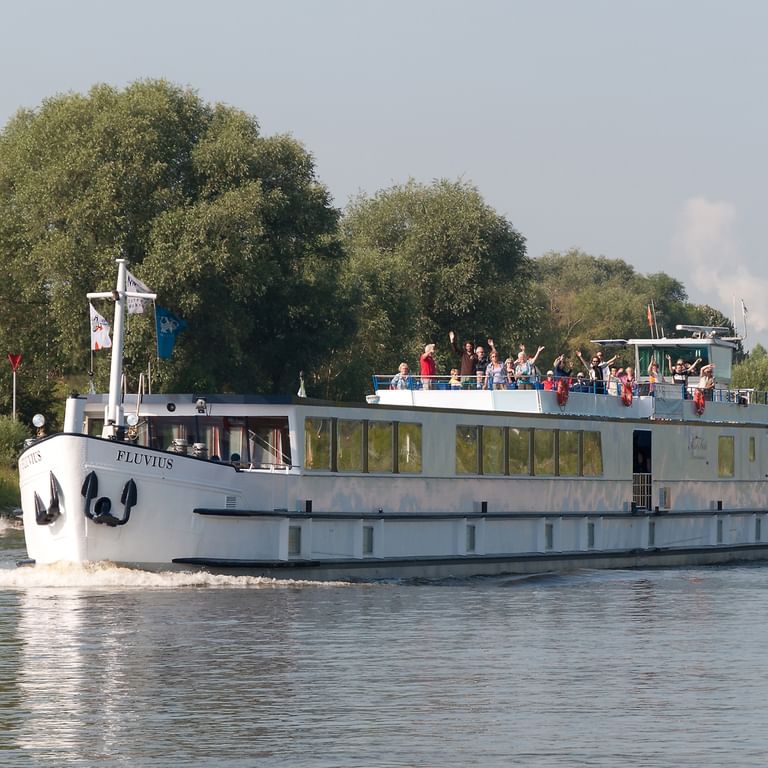 Navire à passagers blanc MS Fluvius naviguant sur une rivière avec des passagers sur le pont, entouré d'arbres verts le long de la rive.